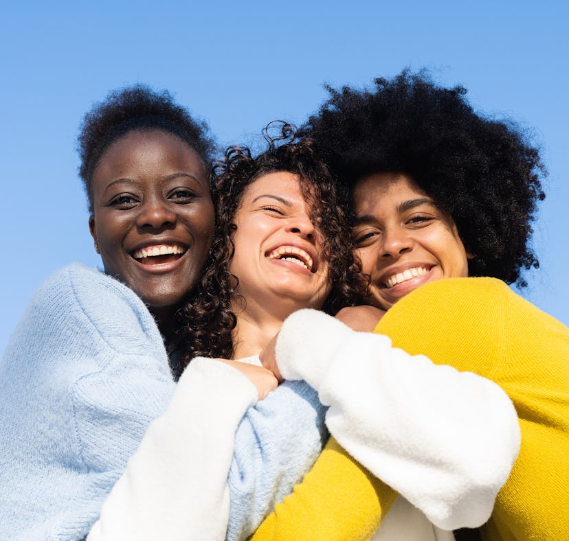 Three cheerful multi-ethnic young women embracing on blue background and two of them looking at the ...