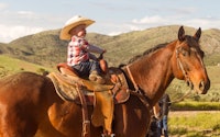 A young child wearing a cowboy hat, riding a horse in a picturesque country setting in an article ab...