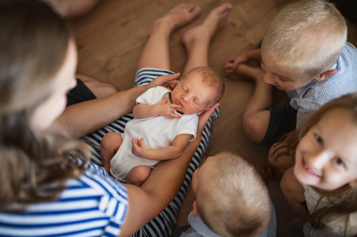 Young mother holding their newborn baby and three small children sitting next to her indoors at home...