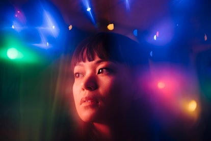 a young woman, surrounded by colorful lights, looks off into the distance as she considers how the f...