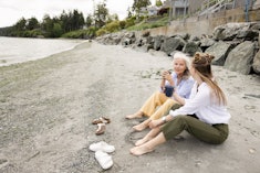 Barefoot mother and daughter bonding, sitting and drinking coffee, talking on rocky beach