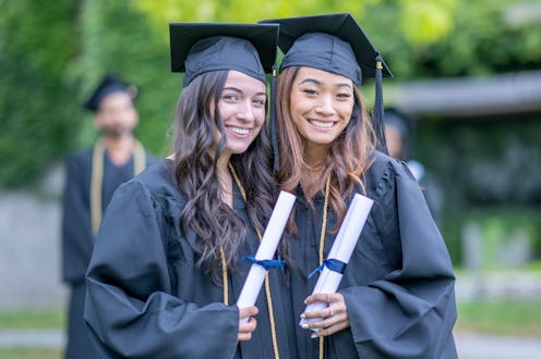 Two female University graduates pose together of a portrait.  They are both wearing formal gowns and...