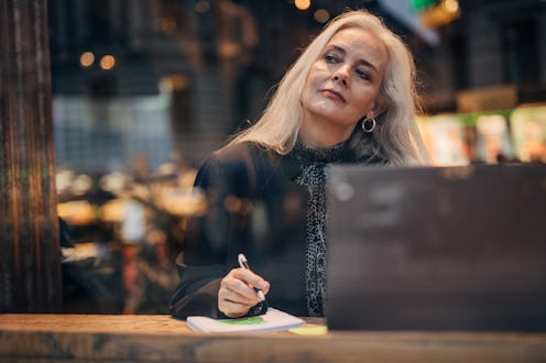 Senior woman using laptop in coffee shop and writing something