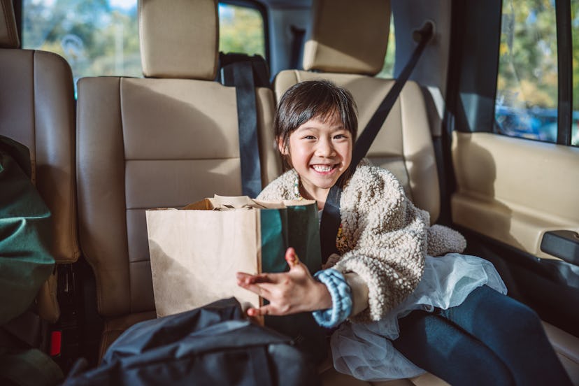 Lovely cheerful girl sitting in car, smiling joyfully at the camera while holding a paper bag of tak...