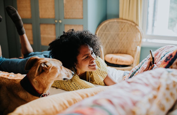 A happy young woman with curly dark hair lounges on the couch with her dog in a sunny, warmly lit ro...