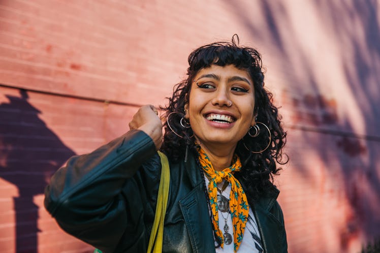A stylish young woman with a big smile and hoop earrings smiles in front of a sunny building while l...