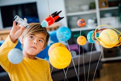 Little boy playing with his homemade planetarium as he holds an astronaut. A rocket hangs above. Arm...