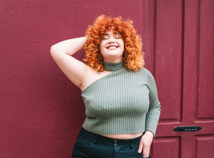 A young woman with curly red hair is seen from the hips up as she smiles with her eyes closed, leani...