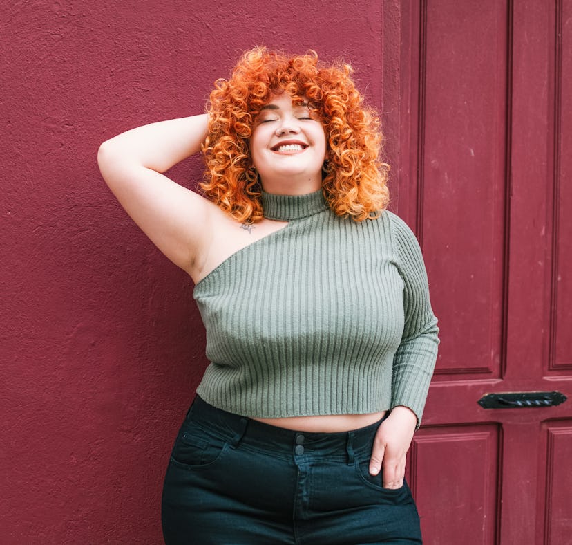 A young woman with curly red hair is seen from the hips up as she smiles with her eyes closed, leani...