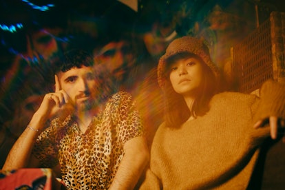A seated young man with a beard in a leopard print shirt and a young woman in a bucket hat pose for ...