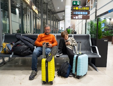 Father and son sitting on a bench using a mobile phone while waiting for flight at the airport loung...