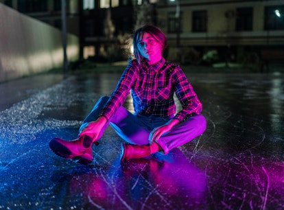 A young woman illuminated by blue and pink light sits on wet, reflective pavement in a basketball co...