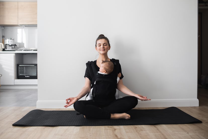 Beautiful young mother with newborn baby in carrier making yoga together at home. Lotus position, Pa...