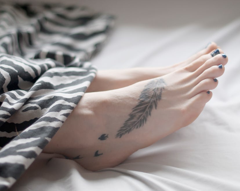 Womans foot amongst bed clothes with a feather tattoo and small birds, in a story about tattoos tatt...