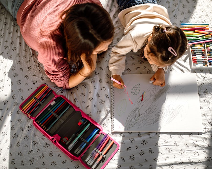 Mother and daughter having fun drawing with coloured pencils in a bedroom, lying down on a bed.