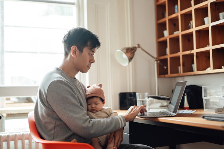 Young Asian father using laptop while holding his baby. Family responsibility. Modern fatherhood.