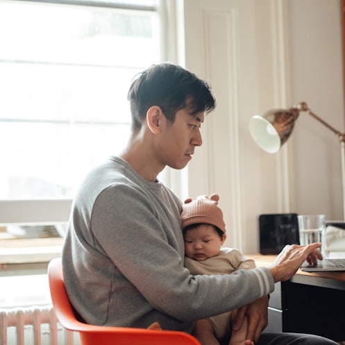 Young Asian father using laptop while holding his baby. Family responsibility. Modern fatherhood.