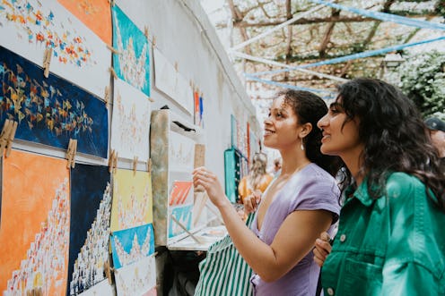 Two woman vacationing together in Europe, stop to admire the paintings at an outdoor market stall.