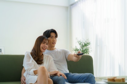 Asian couple cozily watches TV on the sofa. The woman rests her head on the man's shoulder, sharing ...