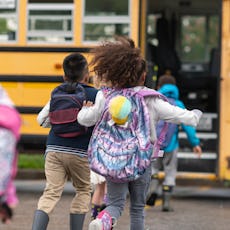 A multi-ethnic group of elementary age children are getting on a school bus. The kids' backs are to ...