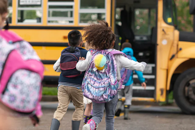 A multi-ethnic group of elementary age children are getting on a school bus. The kids' backs are to ...