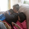 Grandmother comforts her grandson who is crying on the couch at home. Her granddaughter plays on the...