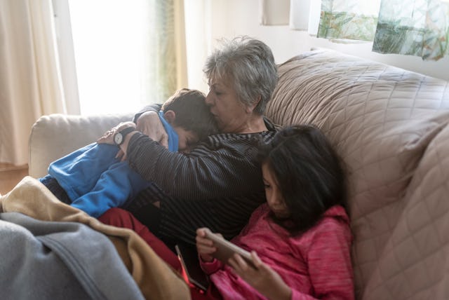 Grandmother comforts her grandson who is crying on the couch at home. Her granddaughter plays on the...