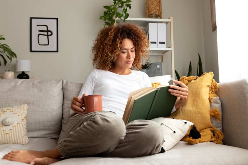 Young multiracial woman reading a book at home, drinking tea sitting on the sofa in cozy living room...