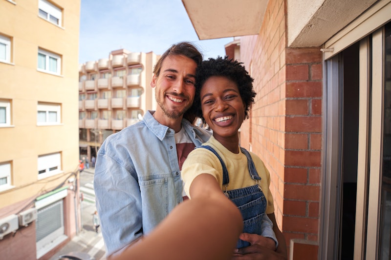 Happy selfie of a multiethnic couple enjoying the sunny day at home in the balcony, looking at camer...