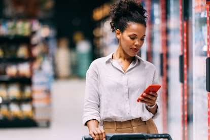 A smiling Latin female holding a cart while using an app on her smartphone at the grocery store.