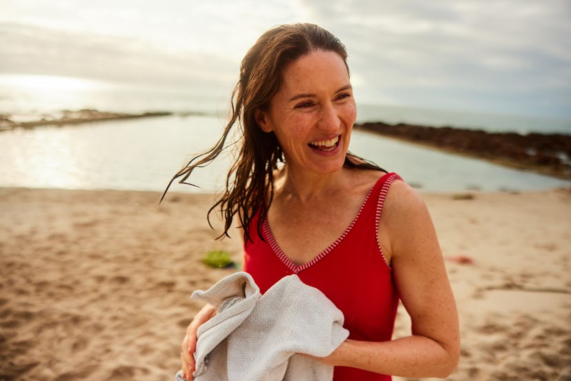 Laughing mature woman wearing a bathing suit drying herself with a towel on a beach after going swim...