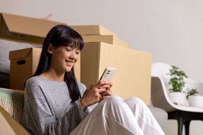 Happy young Asian woman using the Moving App on her phone, surrounded by cardboard boxes and furnitu...