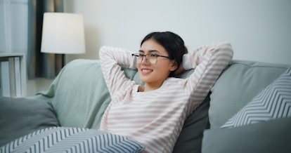 Footage of young Asian woman in glasses sitting on comfortable sofa with her arms to back head while...