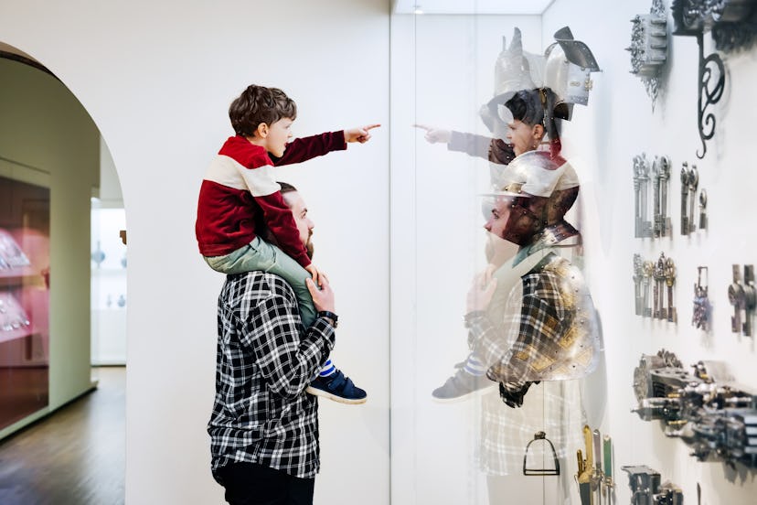 A young boy sitting on his father's shoulders and pointing at an exhibit while visiting a museum tog...