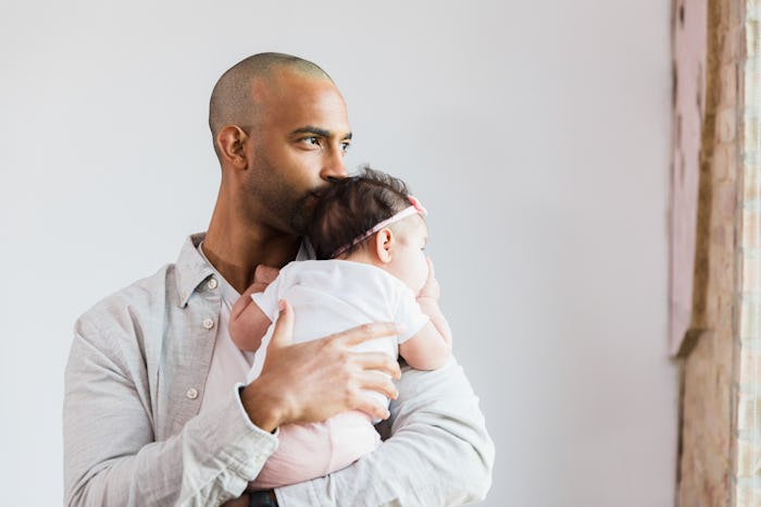 The mid adult stay-at-home father looks out the window in his home as he carries his baby daughter.
