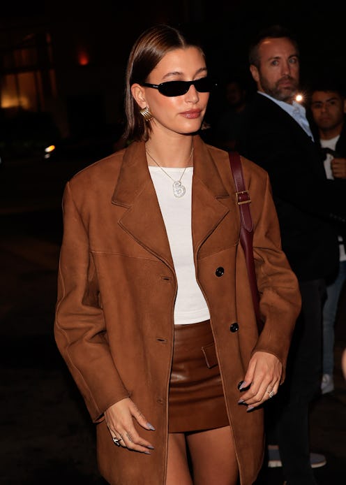 Hailey Bieber wears a brown leather duster coat, a leather skirt, and white tee