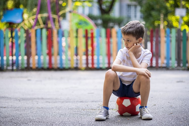 Portrait of lonely kid at the playground sitting on a soccer ball.