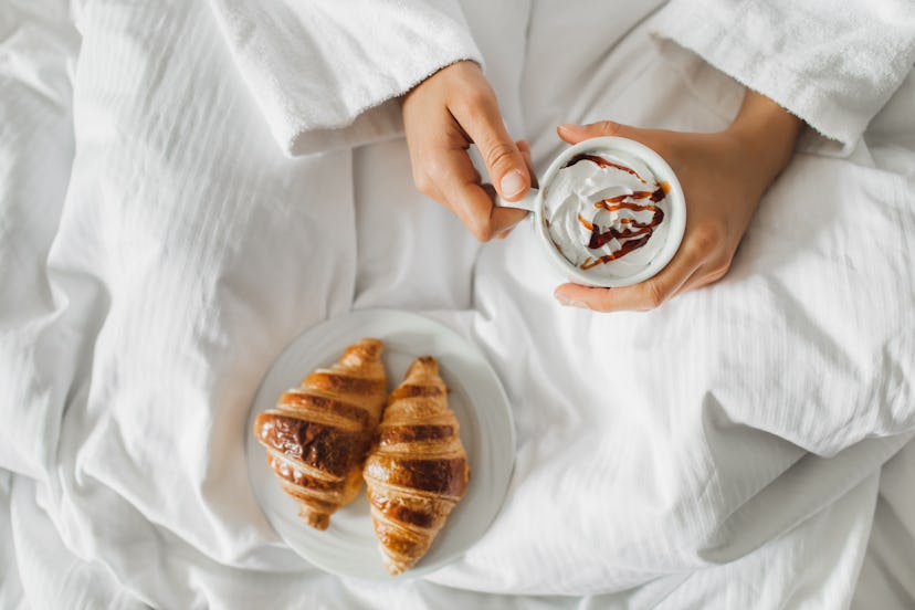 Overhead view of woman hands holding white cup of cappuccino with cream in bed. Two crispy croissant...