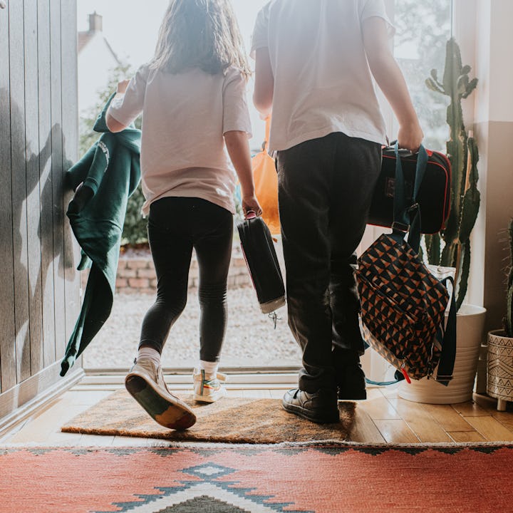 Two young children exit a doorway at the same time, holding backpacks and lunch bags and wearing a s...