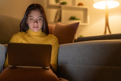 Young woman doing homework on laptop at night, in the living room in her apartment