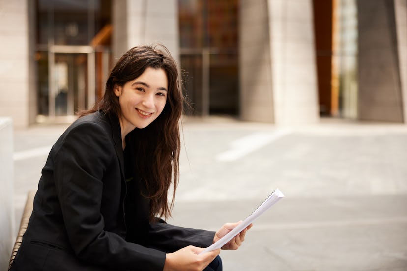 Businesswoman looks up from a document she is reading outdoors on a public bench and smiles at the c...