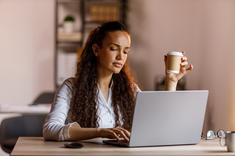 A focused businesswoman using a laptop at home office, surrounded by technology and a cup of coffee.
