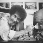 TV painting instructor/artist Bob Ross using a large paint brush to touch up one of his large seasca...