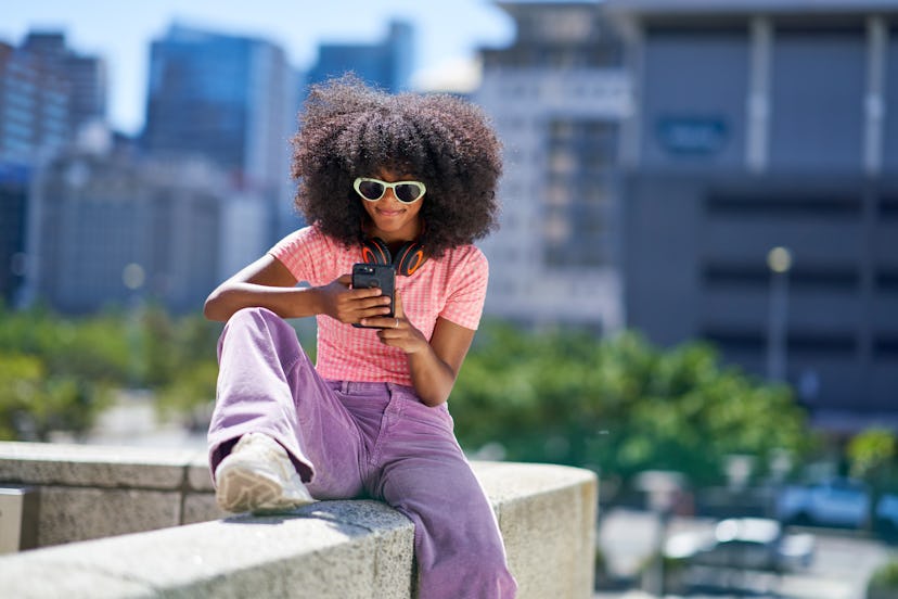 Young woman in sunglasses with headphones using smart phone on ledge in sunny city