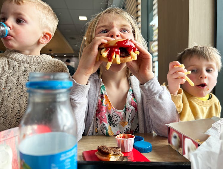 Young girl devours a hamburger loaded with fries and ketchup, while her brothers finish their lunch