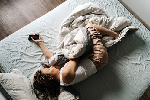 Woman in home clothes lying in the bed with mobile phone. Work at home. Portrait of a dreadlocks bra...