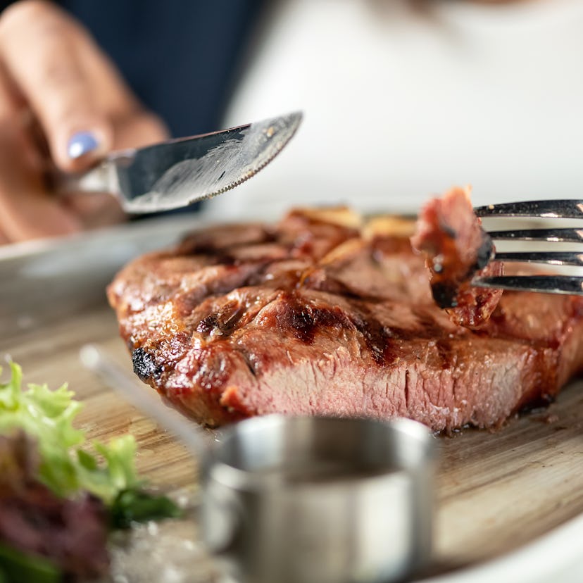 Eating steak. Knife and fork in hands of woman. Pork steak on wooden board. Grilled meat. Lunch at r...