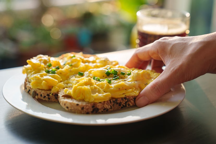 A cropped image of a woman's hand holding a piece of toasted bread with scrambled eggs on top, as sh...