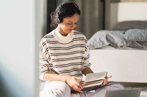A young woman is sitting on the floor at home, studying online. She has her laptop, books and note p...