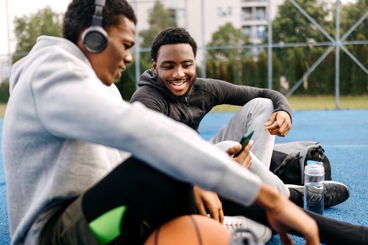 Two young smiling friends using phone while sitting on a ground at outdoor basketball court, listeni...